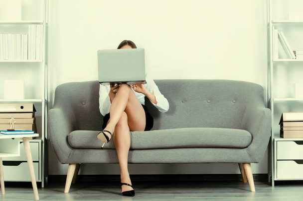 Beautiful secretary legs. Seductive sexy secretary freelancer cover face with laptop computer and sexy legs sit on sofa in office. Sexy business woman. Sensual young girl employee - Φωτογραφία, εικόνα