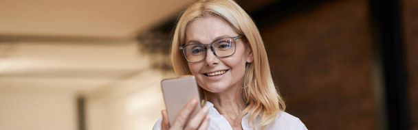 Online Call. Beautiful senior woman in casual wear smiling and talking while making a video call using her smartphone, standing at home. People and communication concept. Horizontal shot - Φωτογραφία, εικόνα