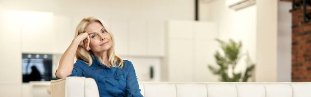 Relaxed mature lady in casual wear looking at camera while sitting on a couch in modern apartment on a daytime. People, business, career concept - Photo, Image