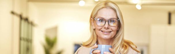 Beautiful mature businesswoman in glasses smiling at camera and drinking coffee while working on the laptop in office. People, business, career concept. Front view - Photo, Image