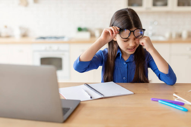 Augenprobleme. Müdes Schulmädchen reibt sich schmerzende Augen bei den Hausaufgaben am Laptop, leidet unter dem Ermüdungs- und Trockenheitssyndrom und trägt zu Hause eine Brille am Schreibtisch. Probleme mit der Sehkraft von Kindern - Foto, Bild
