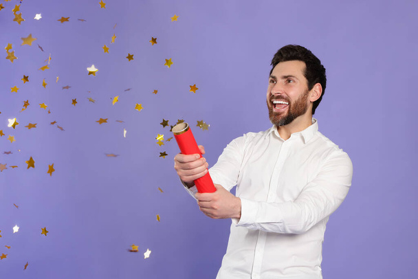 Happy man blowing up party popper on violet background - Zdjęcie, obraz