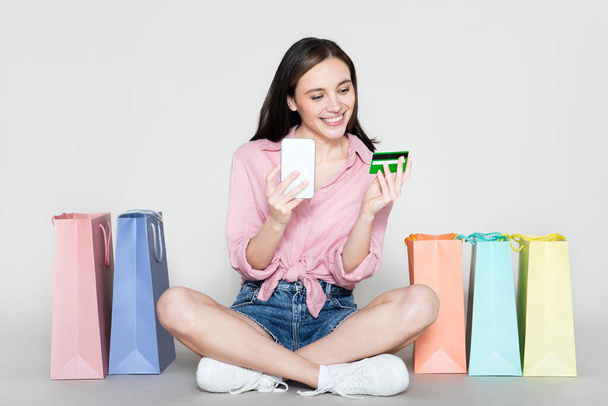 Cool stylish happy smiling young summer woman wearing casual outfit sitting on floor among colorful shopping bags, smartphone, bank card, posing over studio background. E-commerce, retail concept - Zdjęcie, obraz