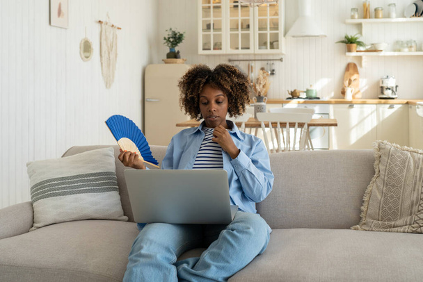 疲れ切ったアフリカ系アメリカ人の女性が青いファンを振って、自宅のノートパソコンで仕事をするのに苦労しています。エアコンのない現代的なアパートで高温の過熱、暑い夏の天気. - 写真・画像
