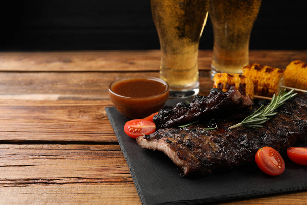 Glasses of beer, tasty fried ribs, grilled corn and sauce on wooden table. Space for text - Photo, image
