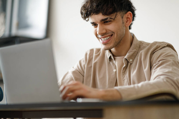 Portrait Of Happy Young Man Using Laptop Working And Learning Online Sitting At Desk Indoors. Middle Eastern Male Manager Typing On Computer And Smiling Posing At Workplace. Selective Focus - Photo, Image