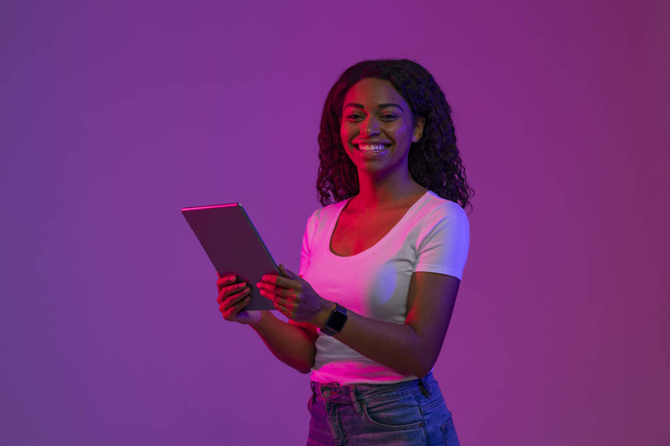 Digitale Freizeit. Glückliche schwarze Frau mit Tablet-Computer in den Händen posiert in Neonlicht über lila Hintergrund, lächelnde Afroamerikanerin genießt moderne Technologien, Kopierraum - Foto, Bild