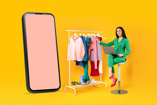 Happy female shopaholic sitting near big cellphone and clothing rail, choosing and buying clothes online, mockup, yellow studio background - Photo, Image