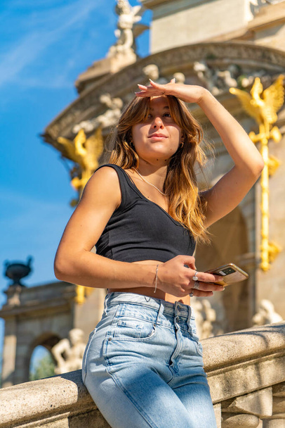 Bela jovem bronzeada em um top preto e jeans e cabelo segurando seu telefone celular e cobrindo os olhos do sol à procura de alguém descansando em uma cerca de escadas de granito - Foto, Imagem