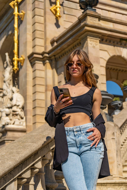 Молодая, загорелая, красивая женщина в черном топе и джинсах в солнцезащитных очках и длинных волосах, смотрит, читает, печатает на мобильном телефоне, стоя на лестнице парка Цитадели - Фото, изображение