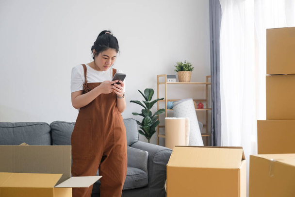 Jonge alleenstaande vrouw met behulp van haar mobiele telefoon te controleren lijst spullen en contract met verhuisbedrijf. - Foto, afbeelding
