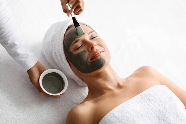 Anti-Aging Skincare. Cosmetologist applying clay mask on face of middle aged woman, beautiful mature female with towel on head lying on table, enjoying beauty tratments in spa salon, closeup - Foto, Imagen