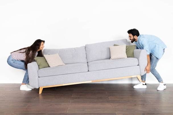 Счастливая возбужденная красивая индийская молодая пара двигает серый диван в комнате над белым фоном стены в новом доме, в полный рост, пространство для копирования. Переезд, переезд на новую квартиру - Фото, изображение