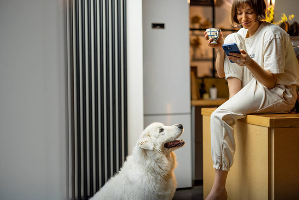 Jeune femme utilise smartphone et boit un café tout en étant assis avec son chien sur la cuisine à la maison. Concept de routine matinale et mode de vie domestique - Photo, image