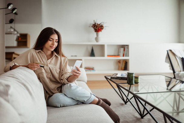Positive millennial european lady shopaholic with credit card looking at phone, order purchases in minimalist living room interior. App for online banking, shopping, sale and cashback - 写真・画像