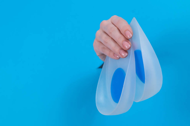 Woman holding orthopedic silicone heel pads. A womans hand sticks out of a hole in a cardboard blue background - Φωτογραφία, εικόνα