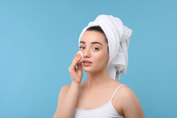 Beautiful woman in terry towel removing makeup with cotton pad on light blue background, space for text - Foto, Imagem