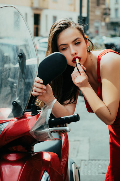 Portrait of beautiful sexy fashion girl model with red lips put lipstick looking in a mirror driving car. Blonde red lipstick looking in the mirror on a moped. face of an attractive brunette woman in - Фото, изображение