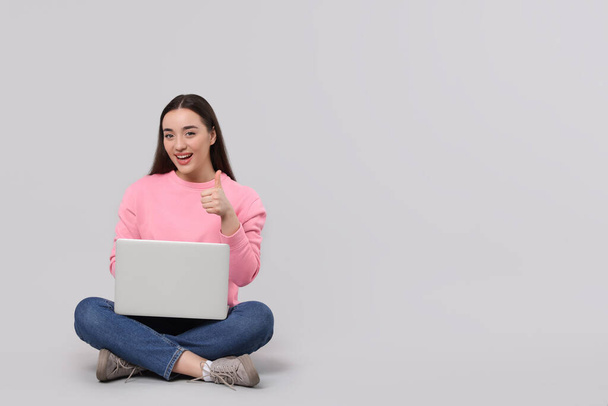 Smiling young woman with laptop showing thumbs up on grey background, space for text - Foto, afbeelding