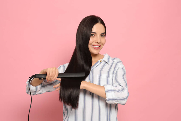 Beautiful happy woman using hair iron on pink background - Foto, afbeelding