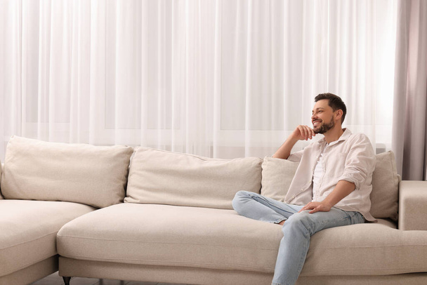 Happy man on resting sofa near window with beautiful curtains in living room. Space for text - 写真・画像