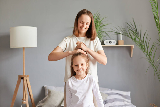 Positive cheerful mother making hairstyle to her daughter while they sitting on bed in bedroom, doing ponytail to kid before going to school in the morning. - Foto, immagini