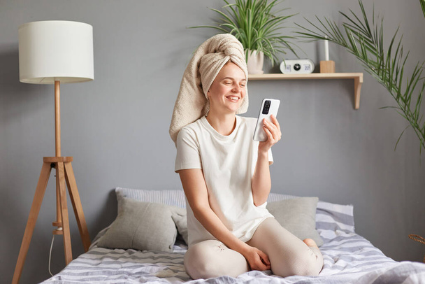 Huomenta, kauno Terve rento nainen istuu sängyllä älypuhelimella, levätä kylpyhuoneen jälkeen, ottaa videopuhelu nauraa ilmaista positiivisia tunteita. - Valokuva, kuva