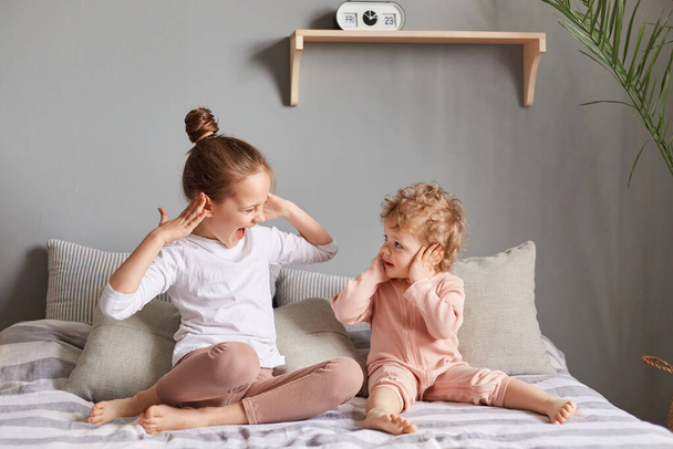 Indoor shot of crazy happy little girls playing in bedroom, two sisters on bed covering ears, wearing home clothing play together expressing happiness. - Foto, afbeelding