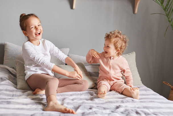 Extremely happy little girls playing in bedroom, laughing loud, being in good playful mood, wearing home clothing, wake up weekend morning and having fun. - Foto, Imagem