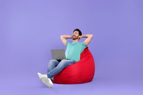 Happy man with laptop sitting on beanbag chair against purple background - Foto, imagen