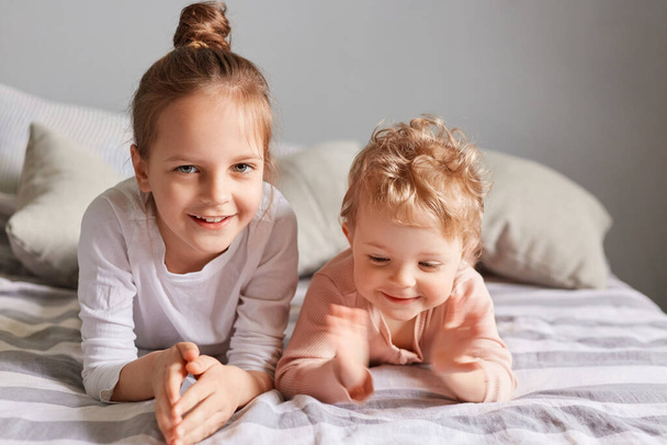 Smiling pretty small kids lying in bed with high spirit enjoying weekend happy little girls playing in bedroom elder sister taking care of infant baby. - Фото, изображение