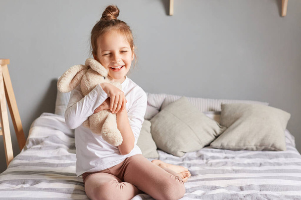 Adorable little girl in home clothing playing with fluffy rabbit , kid having fun with soft toy, child playing with hare embracing favorite bunny smiling happily. - Фото, изображение