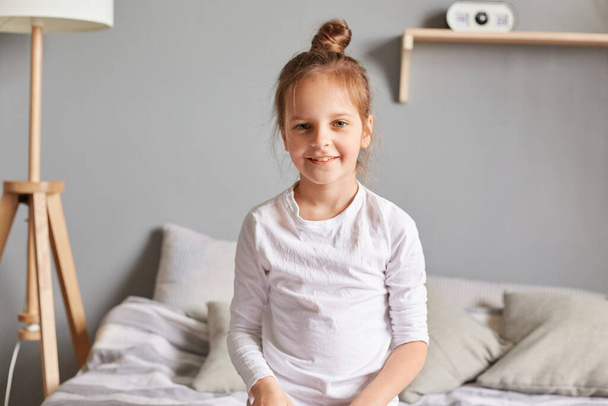 Positive little girl sitting on bed, smiling kid with bun hairstyle wearing white home clothing looking at camera with optimistic expression, cute emotional child. - Photo, image