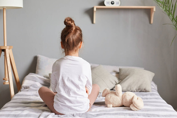 Adorable brown haired girl sitting on bed at bedroom, posing backwards wearing white home clothing, kid with hair bun alone sits on bed. - Zdjęcie, obraz