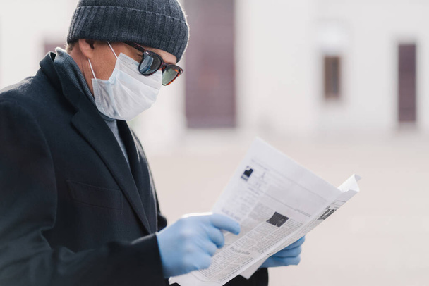 Sepelvaltimoviruksen karanteeni. Vaakasuora kuva aikuisesta miehestä käyttää suojaavaa naamiota ja kertakäyttökäsineitä, lukee tuoretta sanomalehteä, saa selville uutisia pandemiatilanteesta. Infektioiden ehkäisyn käsite
 - Valokuva, kuva