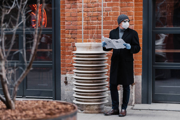 Horizontal shot of young man looks aside, wears medical rubber gloves and face mask, looks aside, holds opened newspaper, reads news, thinks about virus protection. Coronavirus outbreak concept - Foto, afbeelding