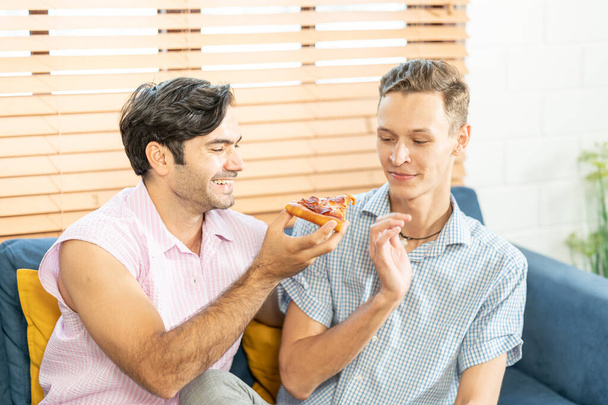 ソファに腰掛け、自宅のリビングルームでビールとピザを飲みながらテレビを見ているカジュアルな服装の2人の男性。LGBTゲイ関係の概念. - 写真・画像