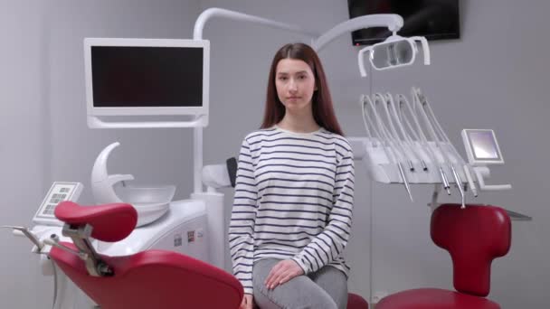 Mosolygó boldog lány mutató hüvelykujj fel gesztus ülni a fogorvosi szék beltéri szekrény vár stomatológus a szájsebészeti beavatkozás Egészségügyi fogszuvasodás zománc kezelés. - Felvétel, videó