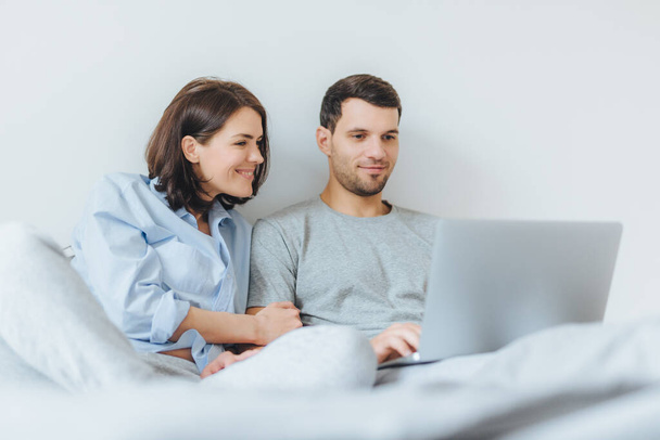 Affectionate female and male have joy together, watch film in bedroom on laptop computer, enjoy togetherness and free internet connection. Lovely couple in love have good time in bedroom - Foto, afbeelding