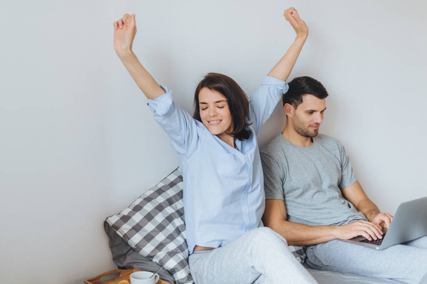 Pretty female stretches as just awoke, has good morning, enjoys day off and her husband sits near in bed, plays computer games on laptop, being concentrated. Family, leisure and home concept - 写真・画像