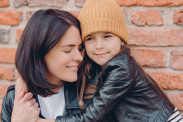 Close up shot of tender pleased young lady smiles gladfully and closes eyes, recieves hug from small daughter who wears leather jackt and hat, pose together over brick wall. Motherhood concept - Фото, изображение