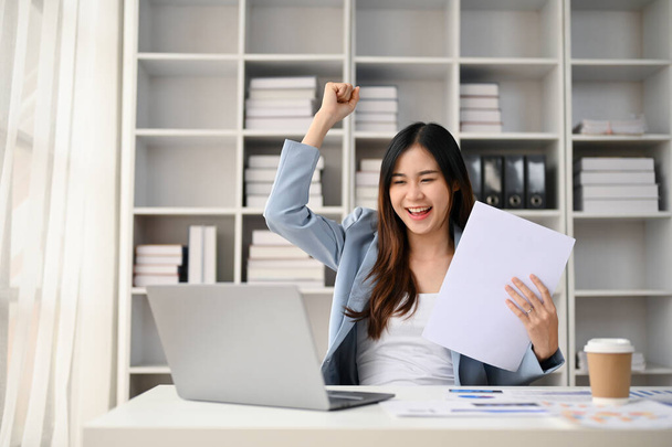 Fröhliche asiatische Millennial-Geschäftsfrau blickt auf ihren Laptop-Bildschirm, hebt die Hand, jubelt und feiert ihren Projekterfolg an ihrem Schreibtisch im Büro. - Foto, Bild