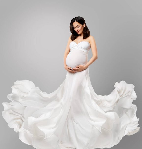 Ciężarna kobieta w białej długiej sukience latająca na wietrze patrząca na brzuch. Piękna matka w jedwabnej sukni falującej na szarym tle. Ubrania ciążowe. Opieka zdrowotna nad matkami - Zdjęcie, obraz