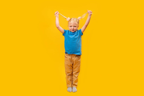 Smiling preschooler wears blue T-shirt on yellow background. Full-length portrait of little blond girl with two ponytails. - Fotó, kép