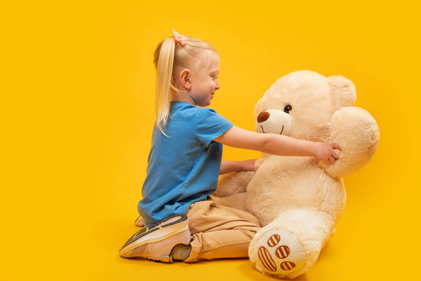 Studio portrait of little girl with large teddy bear on yellow background. Child plays with soft toy - Photo, image