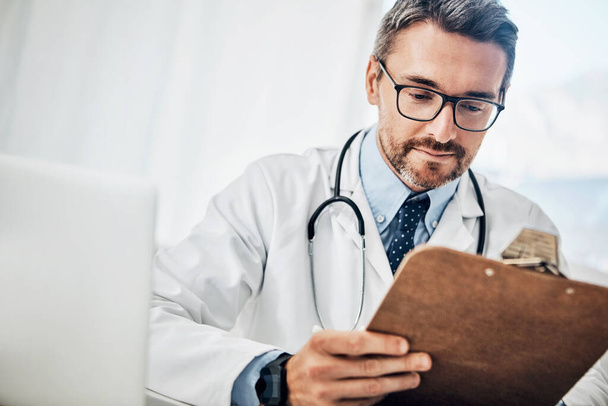 Seinen Patienten durch Krankenakten kennen lernen. ein fokussierter Arzt betrachtet Papierkram auf einem Klemmbrett, während er an seinem Schreibtisch sitzt - Foto, Bild