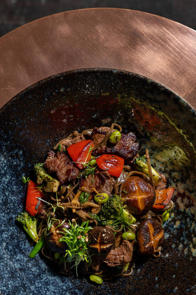Soba noodles with veal and mushrooms, paprika, broccoli and microgreen sprouts in a dark ceramic plate. The plate is on a copper tabletop. - Photo, Image