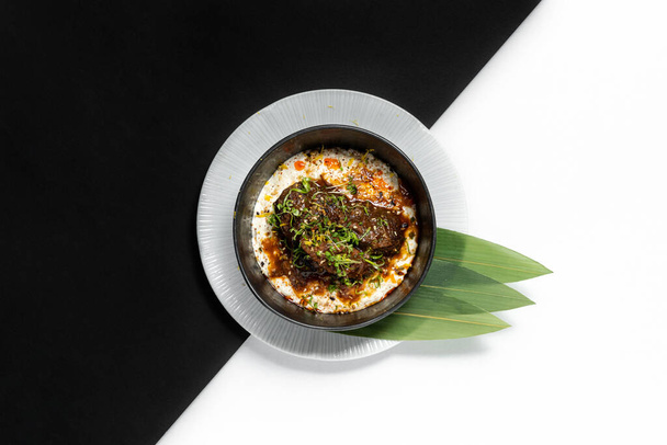 Kalfskaken in unagi saus met groene uien bovenop aardappelpuree. Het eten ligt in een donkere keramische kom, op een lichtplaat met bamboe bladeren. De gerechten staan op een papieren zwart-wit ondergrond. - Foto, afbeelding