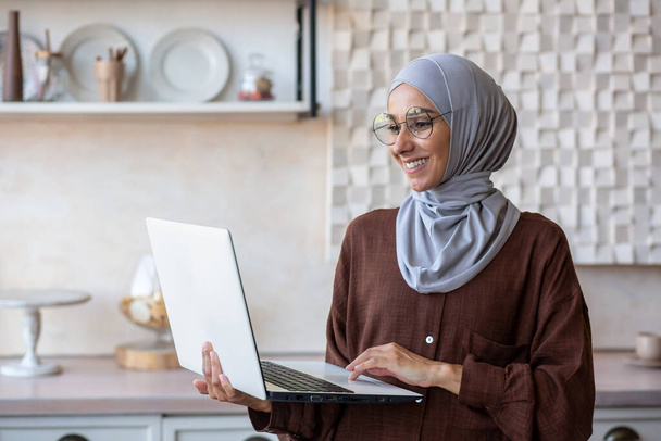 Mujer de negocios alegre y exitosa en hijab en casa usando el ordenador portátil mientras está de pie, mujer musulmana trabajando remotamente. - Foto, imagen