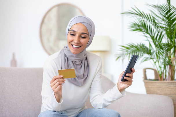 Νεαρή όμορφη Ασιάτισσα γυναίκα με μαντίλα κάθεται στον καναπέ στο σπίτι, μουσουλμάνα γυναίκα που κατέχουν τραπεζική πιστωτική κάρτα και τηλέφωνο στο χέρι, ευτυχώς ψώνια σε απευθείας σύνδεση στο ηλεκτρονικό κατάστημα. - Φωτογραφία, εικόνα